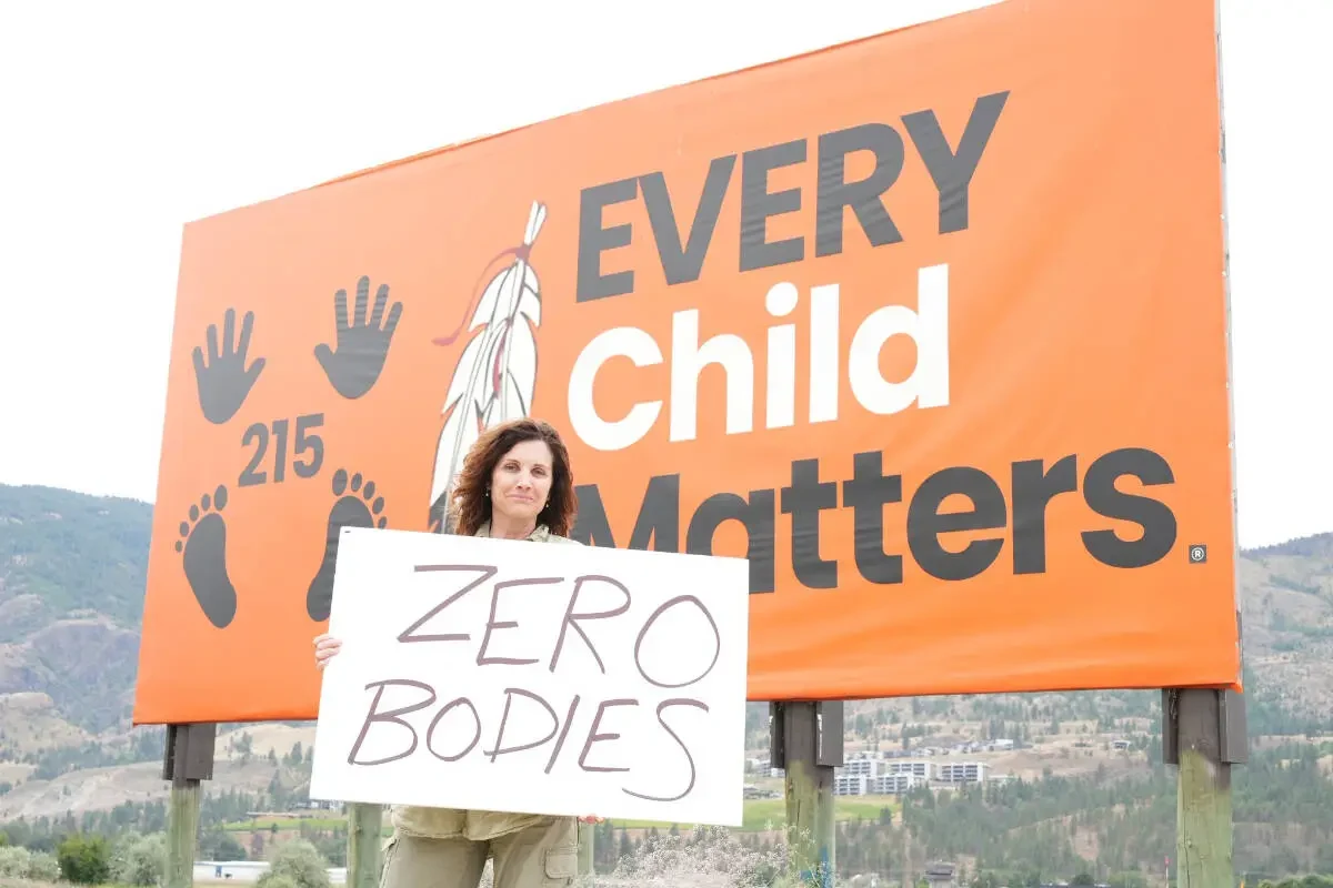 Dallas Brodie standing in front of an 'Every Child Matters' billboard on Penticton Indian Band land, holding a handwritten sign that reads 'Zero bodies'.